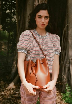 Bolso de cuero marrón tipo bucket con una abertura ajustada, que exhibe una textura suave y un diseño minimalista, sostenido con las manos frente a un fondo natural.