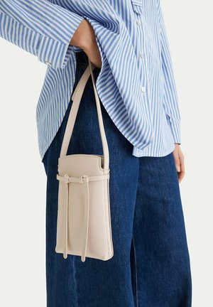 Osoba v modrém pruhovaném tričku a tmavých džínách držící malou béžovou koženou kabelku přes rameno za popruh.