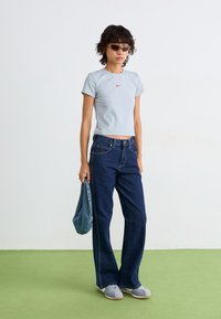 Kvinne iført en lys grå Nike crop t-skjorte, mørkeblå jeans, grå joggesko, solbriller, holder en blå denimveske, stående på grønn gulv.