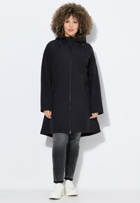 Veste à capuche noire avec une fermeture éclair intégrale, dotée d'une couleur intérieure contrastante, de poches latérales et d'une coupe longue et décontractée. Associée à un jean gris et des baskets blanches.
