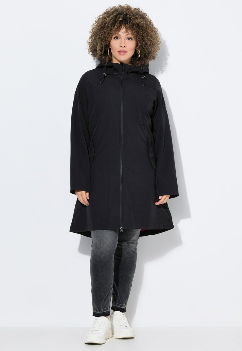 Veste à capuche noire avec une fermeture éclair intégrale, dotée d'une couleur intérieure contrastante, de poches latérales et d'une coupe longue et décontractée. Associée à un jean gris et des baskets blanches.