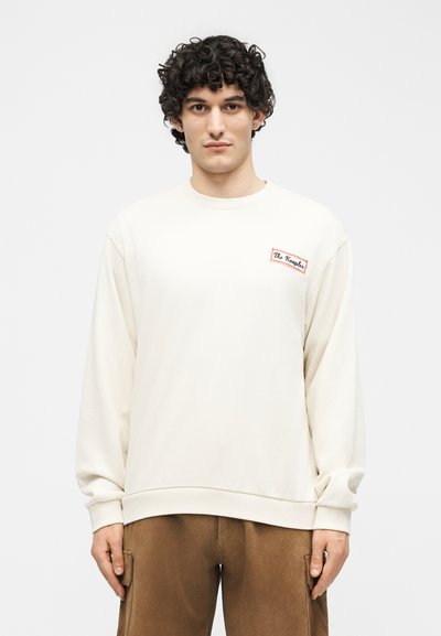 Νεαρός άνδρας με σγουρά σκούρα μαλλιά φοράει λευκό φούτερ με μικρό λογότυπο στο στήθος και καφέ παντελόνι cargo, στέκεται μπροστά σε απλό φόντο.