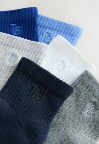 Six paires de chaussettes côtelées en bleu, bleu clair, blanc, gris, marine et gris clair, chacune avec un petit logo brodé "TB".