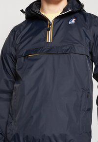 Chaqueta impermeable azul marino con media cremallera, detalles de franjas reflectantes y un bolsillo delantero con cremallera. Cuenta con capucha y cordón ajustable.