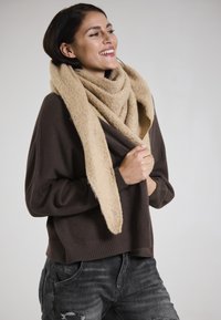 Brown knitted sweater with a loose fit, paired with a chunky beige scarf. The model's outfit includes distressed dark jeans.