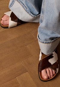 Bruine en witte koeienhuid sandalen met een kruisstropontwerp. De zool is donker en de banden hebben een zachte, gestructureerde afwerking.