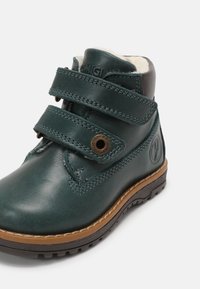 Bota de tobillo de cuero verde oscuro con dos correas de velcro, suela de goma marrón y detalles de costura. Forrada con material suave para mayor comodidad.