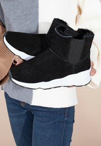Botas de tobillo de ante negro con suelas de goma blanca, paneles laterales elásticos y un forro interior suave, sostenidas por una persona que lleva un suéter multicolor.