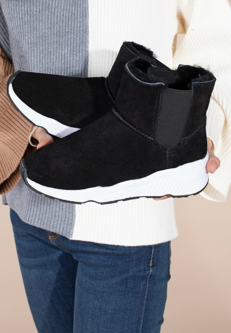 Botas de tobillo de ante negro con suelas de goma blanca, paneles laterales elásticos y un forro interior suave, sostenidas por una persona que lleva un suéter multicolor.