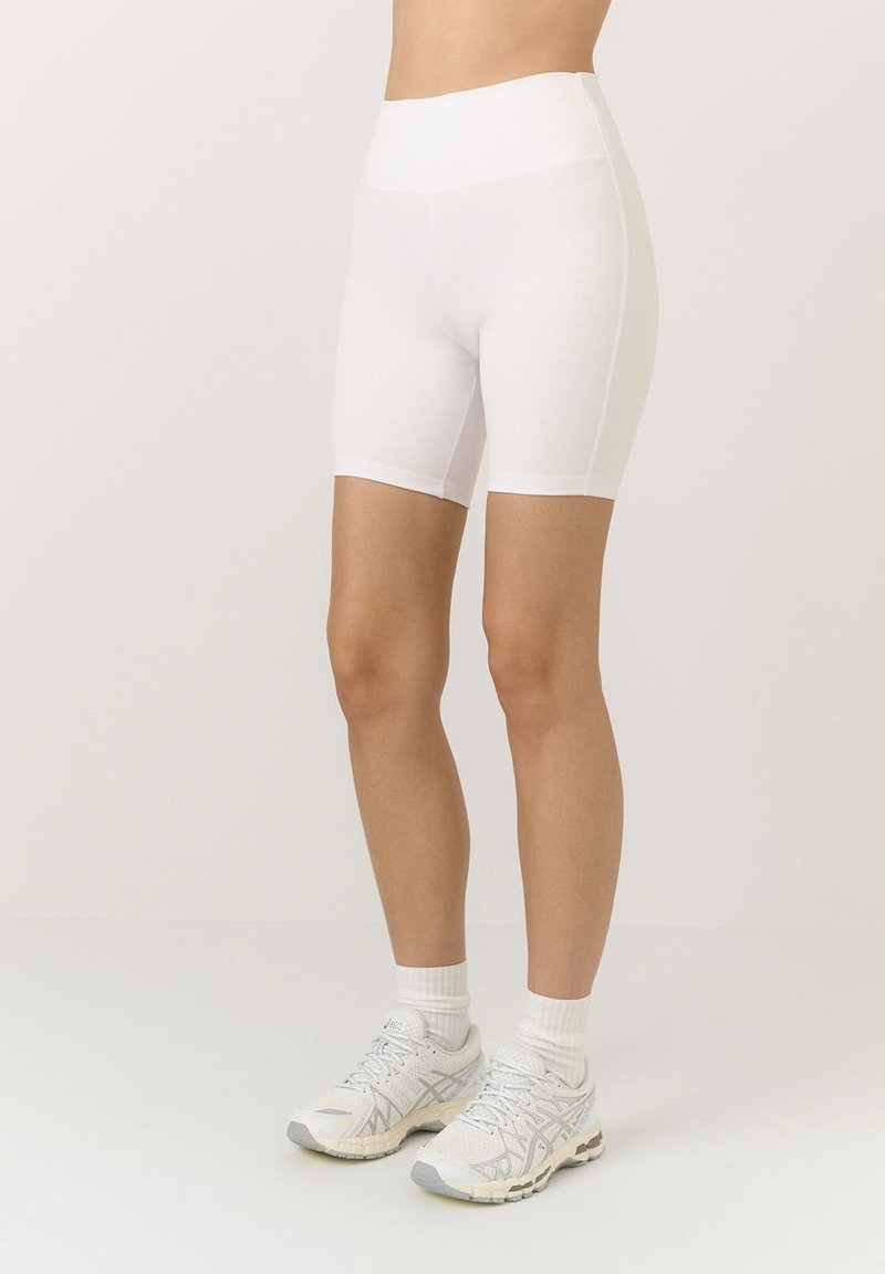 Białe szorty z wysokim stanem o gładkiej, elastycznej teksturze, sięgające powyżej połowy uda. Noszone z jasnymi butami sportowymi i skarpetkami.