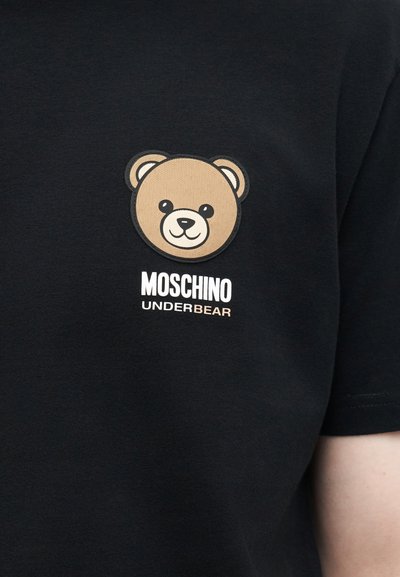Χαμογελαστό καρτούν πρόσωπο αρκούδας πάνω από λευκό κείμενο «MOSCHINO» και μπεζ κείμενο «UNDERBEAR» σε μαύρο ύφασμα, μέρος ενός κοντομάνικου πουκάμισου.