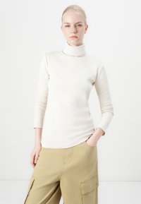 Tejfehér, garbós felső hosszú ujjakkal, bordás anyagból készült. Formázott dizájn, khaki cargo nadrággal viselve. Minimalista stílus.