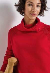 Roter Strickpullover mit einem Stehkragen, der strukturierte Akzente und zwei Knöpfe am Kragen aufweist. Weiches Material mit gerippten Bündchen.