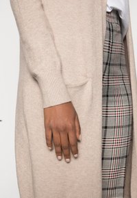 Beige langer Cardigan aus weichem Material, mit gerippten Bündchen und einer Fronttasche. Trägt man über karierter Hose mit einem weißen Hemd darunter.