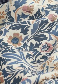 Tissu crème avec un motif floral comprenant des feuilles bleues et des fleurs roses, orange et bleues, présenté en plis doux et en gros plan.