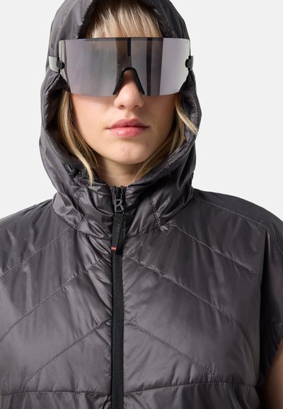 Kvinde iført en grå vadet hættejakke med lynlås og store reflekterende briller med wraparound-stil mod en ensfarvet baggrund.