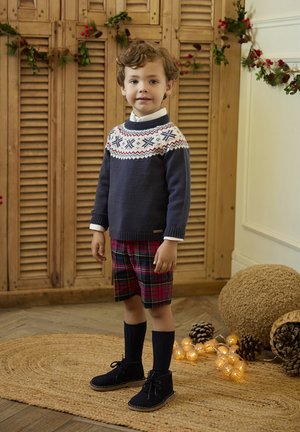 Niño vestido con un suéter azul marino con un cuello decorado, pantalones cortos de cuadros rojos y verdes, calcetas navy hasta la rodilla y zapatos de ante negros.