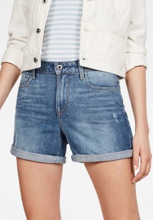 Mujer con pantalones cortos de mezclilla azul enrollados, con bolsillos delanteros, botón y cremallera, combinados con una chaqueta blanca corta y una camiseta a rayas.