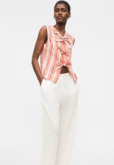 Γυναίκα που φοράει κοραλλί αμάνικη μπλούζα με λευκές ρίγες και τρία φιόγκια μπροστά, καθώς και άνετο λευκό υφασμάτινο παντελόνι, στέκεται με τα χέρια στις τσέπες.