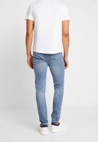 Úzké, vybledlé modré džíny s pětipokotovým designem, nošené s obyčejným bílým tričkem a bílými teniskami, pohled zezadu.