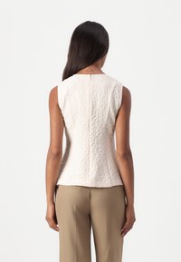 Femme aux longs cheveux foncés portant un haut blanc texturé sans manches et un pantalon beige, debout de dos sur un fond uni.