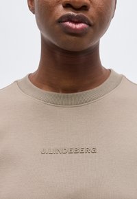 Κοντινή λήψη ατόμου που φοράει μπεζ μπλούζα με στρογγυλή λαιμόκοψη και το ανασηκωμένο λογότυπο "J.LINDEBERG" στο στήθος.