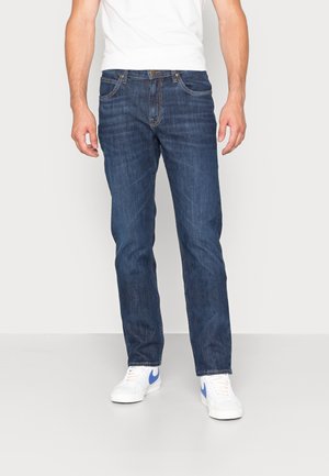 Man draagt donkerblauwe jeans met rechte pijpen, een wit T-shirt met ronde hals en witte sneakers met een blauwe Nike-swoosh, staand op een grijze vloer.