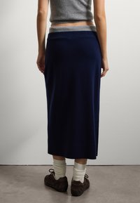 Donna che indossa una gonna midi blu scuro con una cintura grigia, un top senza maniche grigio, calze bianche arricciate e scarpe stringate marrone scuro, in piedi su un pavimento di cemento.