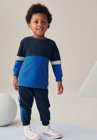 Sinised ja tumesinised pikad triibulised triiksärgid, mida täiendab sobivad tumesinised püksid ja valged tossud, minimalistlik disain.