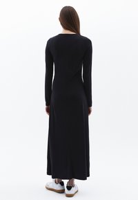 Schwarzes Kleid mit langen Ärmeln, das einen taillierten Oberteil und einen fließenden Rock hat, aus glattem Stoff gefertigt, kombiniert mit weißen Sneakers mit schwarzen Akzenten.