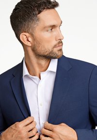 Blazer azul marino con un corte ajustado, que presenta solapas muescas y un único bolsillo en el pecho. Se lleva sobre una camisa blanca de botones con una textura suave.