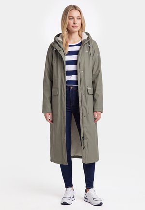 Kvinde iført lang olivengrøn hættejakke, stribet navy og hvid skjorte, mørke jeans og hvide sneakers, stående vendt front.