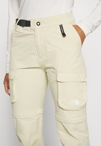 Beige cargobyxor tillverkade av lättviktsmaterial, med ett bälte, flera fickor och The North Face-logotypen på det nedre benet.
