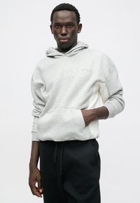 Szara bawełniana bluza z kapturem, z kieszenią typu kangurka, z białą boczną panelą. Mam materiał teksturowany, regulowany kaptur i ściągacze przy rękawach. Założona z czarnymi spodniami.