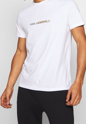Mand iført en hvid kortærmet T-shirt med "KARL LAGERFELD" trykt i metallisk skrift over brystet.