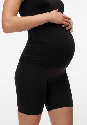 Zwangere persoon draagt zwarte aansluitende shorts en top, houdt zachtjes de buik met beide handen tegen een effen witte achtergrond.
