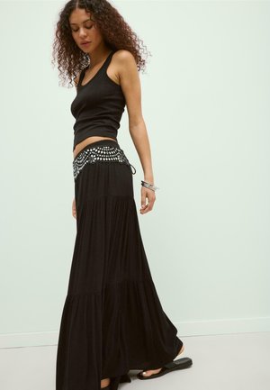 Frau mit lockigem Haar, die ein schwarzes ärmelloses Oberteil und einen langen schwarzen Rock mit dekorativem weißem gemustertem Bund trägt.