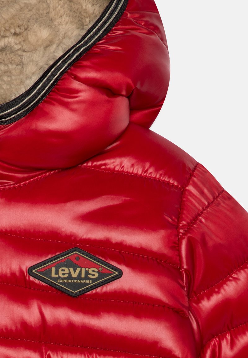 Giacca puffo rossa con finitura lucida, dotata di fodera in pile color cammello, motivo a righe orizzontali quiltate e una toppa del logo Levis nera e oro.