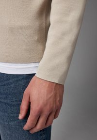 Suéter de punto beige con una silueta ajustada, que presenta una textura suave y puños acanalados, combinado con jeans de mezclilla azul oscuro.