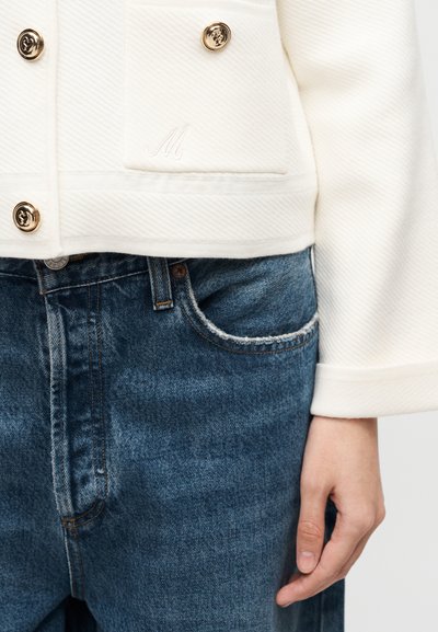 Λευκό υφασμάτινο σακάκι με χρυσά κουμπιά και κεντημένο το γράμμα "M", συνδυασμένο με τζιν ψηλόμεσα μπλε παντελόνια, με το χέρι να ακουμπάει στο πλάι.
