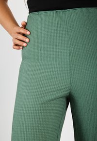 Zelené široké kalhoty s texturou vyrobené z lehkého materiálu s vzorovaným povrchem a vysokým pasem, které drží ruka.