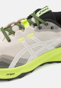 Zapatilla de running Asics con parte superior de malla en beige, talón negro y acentos vibrantes en amarillo. Presenta una suela texturizada marcada como "FF Blast."