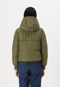 Oliven grønn pufferjakke med hette, quiltet tekstur og elastiske mansjetter, sammen med mørkeblå denimjeans.