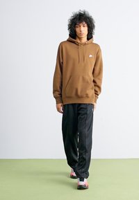 Brun hettegenser med lommer foran og hvit logo, kombinert med svarte bukser. Modellen står på grønt gulv og viser frem sportsfotwear.