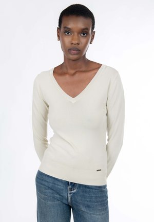 Pull beige clair à manches longues et col en V, fabriqué dans un matériau doux, avec une coupe ajustée et un logo discret à l'ourlet. Porté avec un jean bleu.