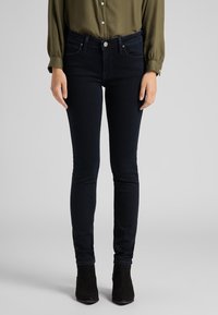 Jeans ajustados de denim azul oscuro con una cintura de altura media, diseño de cinco bolsillos y detalles de costura mínimos, combinados con botas tobilleras.