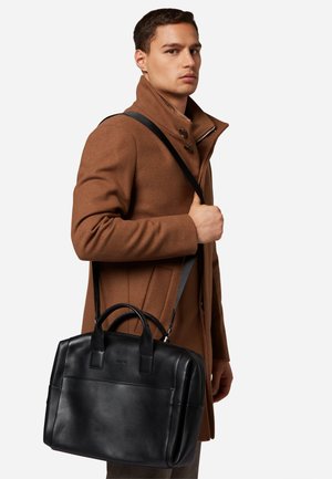 Black leather briefcase with dual handles, detachable shoulder strap, front pocket, and smooth texture, held by a person wearing a brown coat.