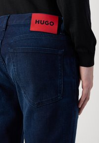 Sötétkék farmernadrág sima textúrával, piros bőr címkével, amelyen feketével van írva: "HUGO", a hátsó derékrészen elhelyezve.