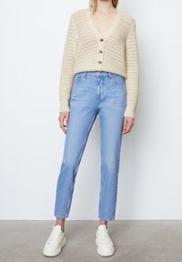 Bežový kardigan s otevřeným pletením, V výstřihem a třemi knoflíky. Světle modré džíny a bílé tenisky s neonovými akcenty doplňují celkový vzhled.