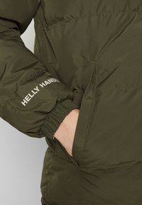 Žalias pūkinis švarkas su elastingomis rankovėmis, šoninėmis kišenėmis ir baltu "HELly HANSEN" logotipu ant rankovės. Lygus, šiltas audinys.